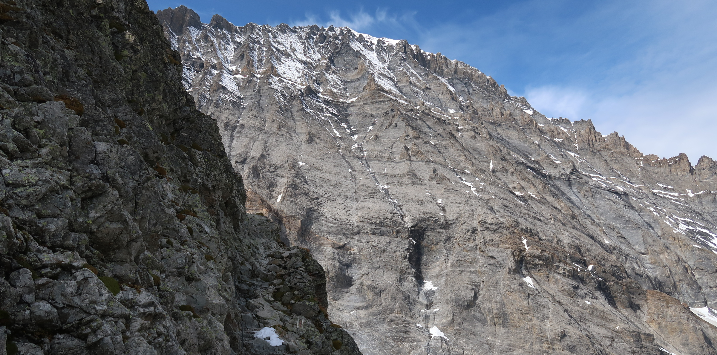 A rocky cliff with a very narrow path cut into it. On one side of the path is a thin cable, in the background a large mountain